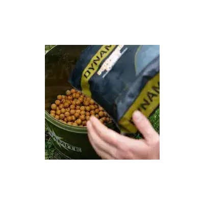 A hand pours golden-brown dynamite baits peppered fruit boilies from a blue and yellow bag into an olive green bucket