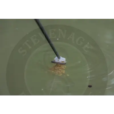 A black fishing rod tip with a white textured bait holder releasing golden pellets into water using tackle guru-fast release pole pots