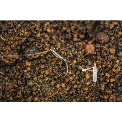 A silver lwg feeder fishing hook with clear plastic swivel and line resting on dark brown gravel from tackle guru
