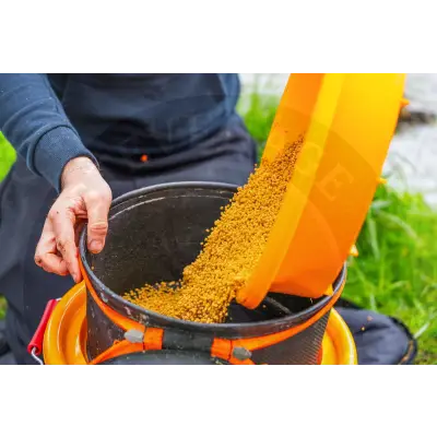 Bright orange plastic scoop pouring golden fish food pellets into black bucket for tackle guru multi riddle use