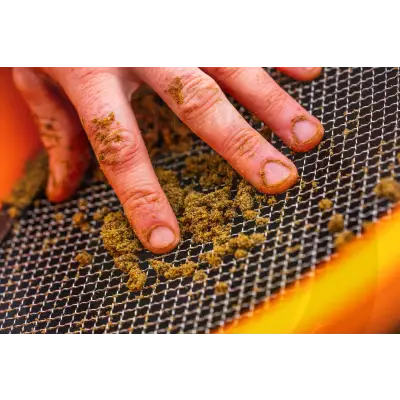 A hand with dirt on fingers rests on a black mesh guru multi riddle screen with brownish-green bait pellets scattered