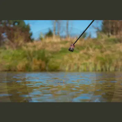 A black fishing rod tip with a dark cylindrical lure near water featuring tackle guru pole pot sprinkle for stevenage bait