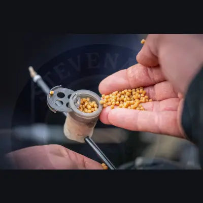 A hand holds a tackle guru pole pot sprinkle lid with black metal lid spilling golden stevenage bait pellets