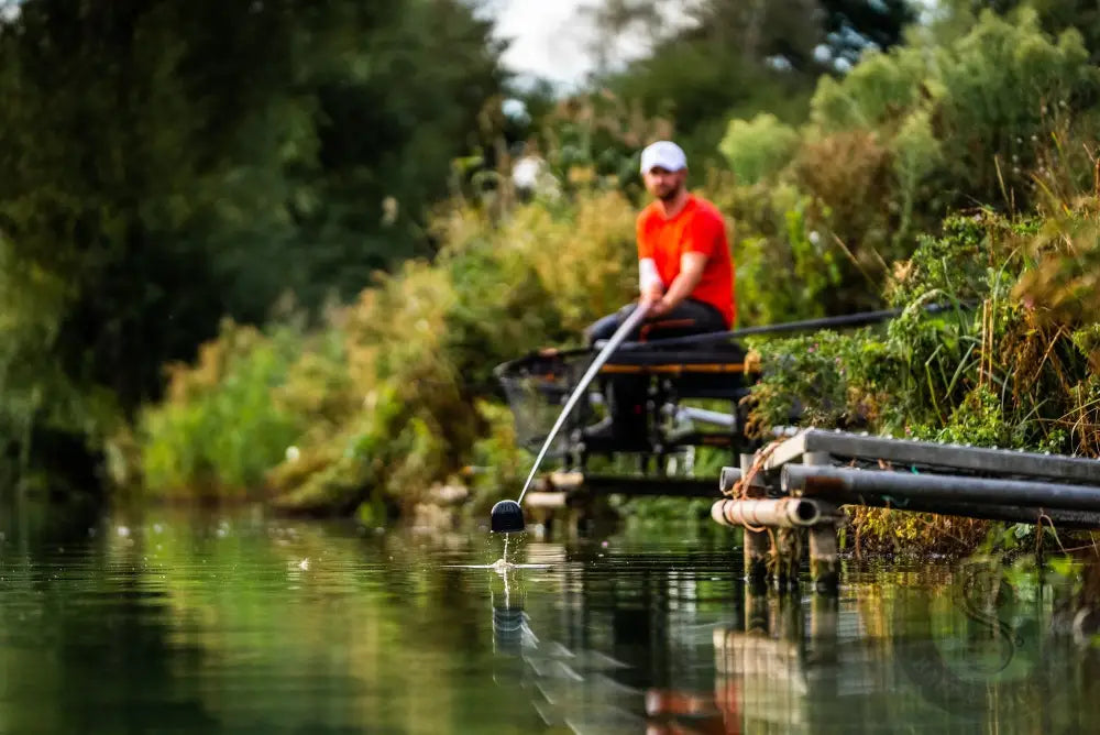 A man in a red shirt and white cap uses rapid release pole cups on a fishing platform holding a black rod with stevenage bait