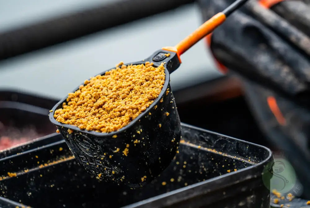 Black plastic measuring scoop with orange handle filled with golden brown fish bait pellets for tackle guru rapid release pole cups