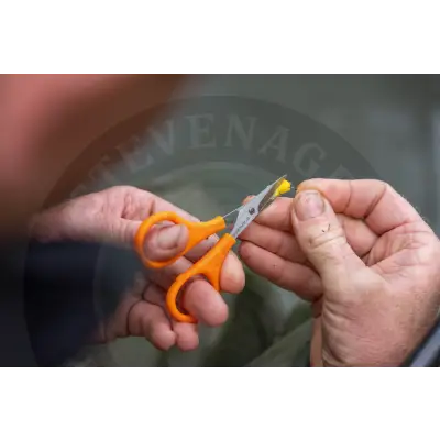 Orange-handled metallic serrated rig scissors from Tackle Guru trimming a small yellow object with fluorescent coloured handles